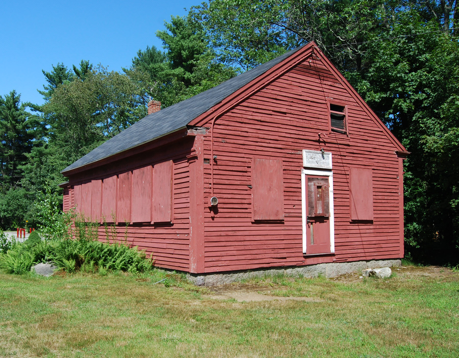 Weathered red wooden schoolhouse with boarded windows surrounded by grass, ferns, and tall trees