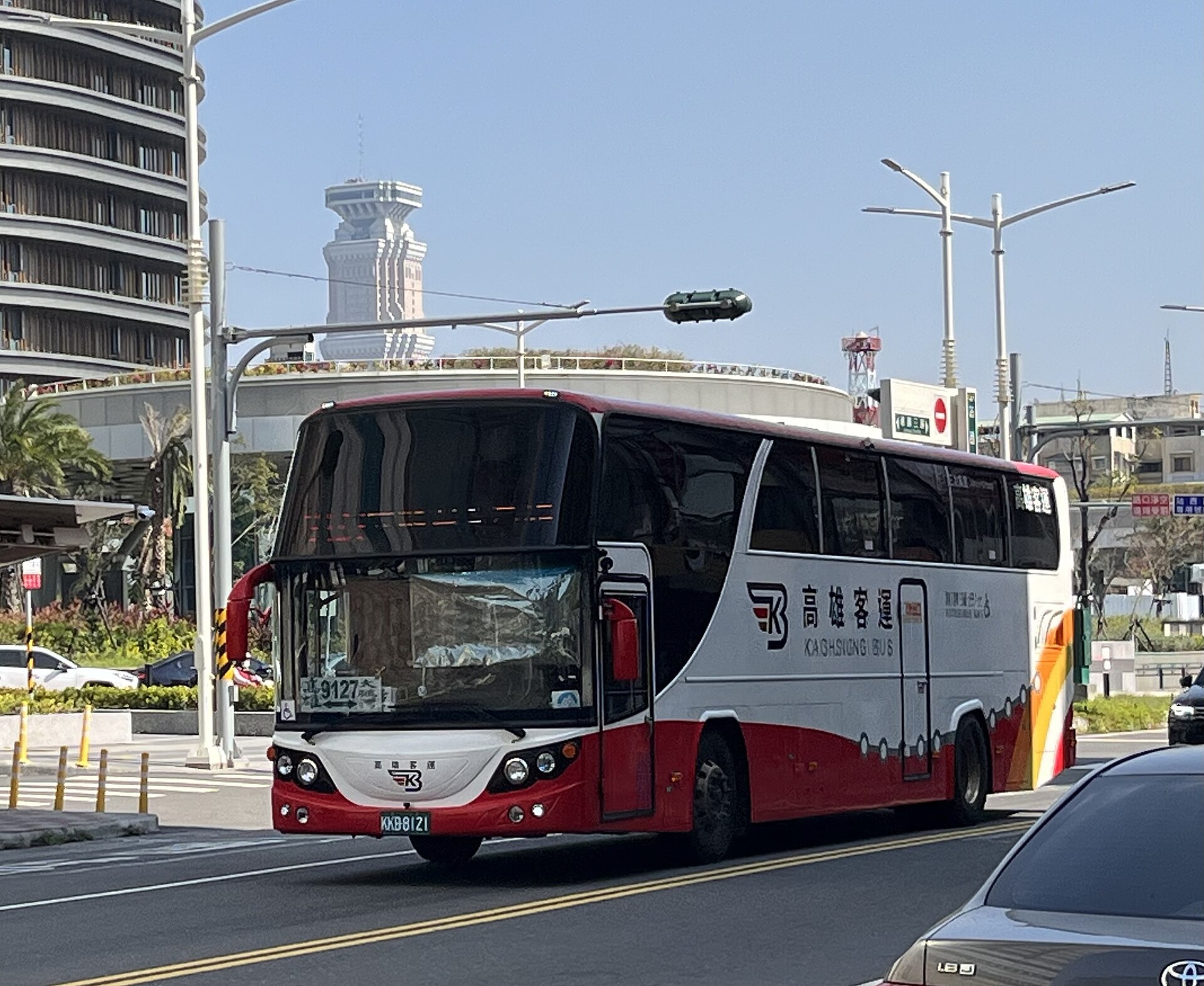 Red, white, and black coach bus traveling through a bright city intersection with modern buildings