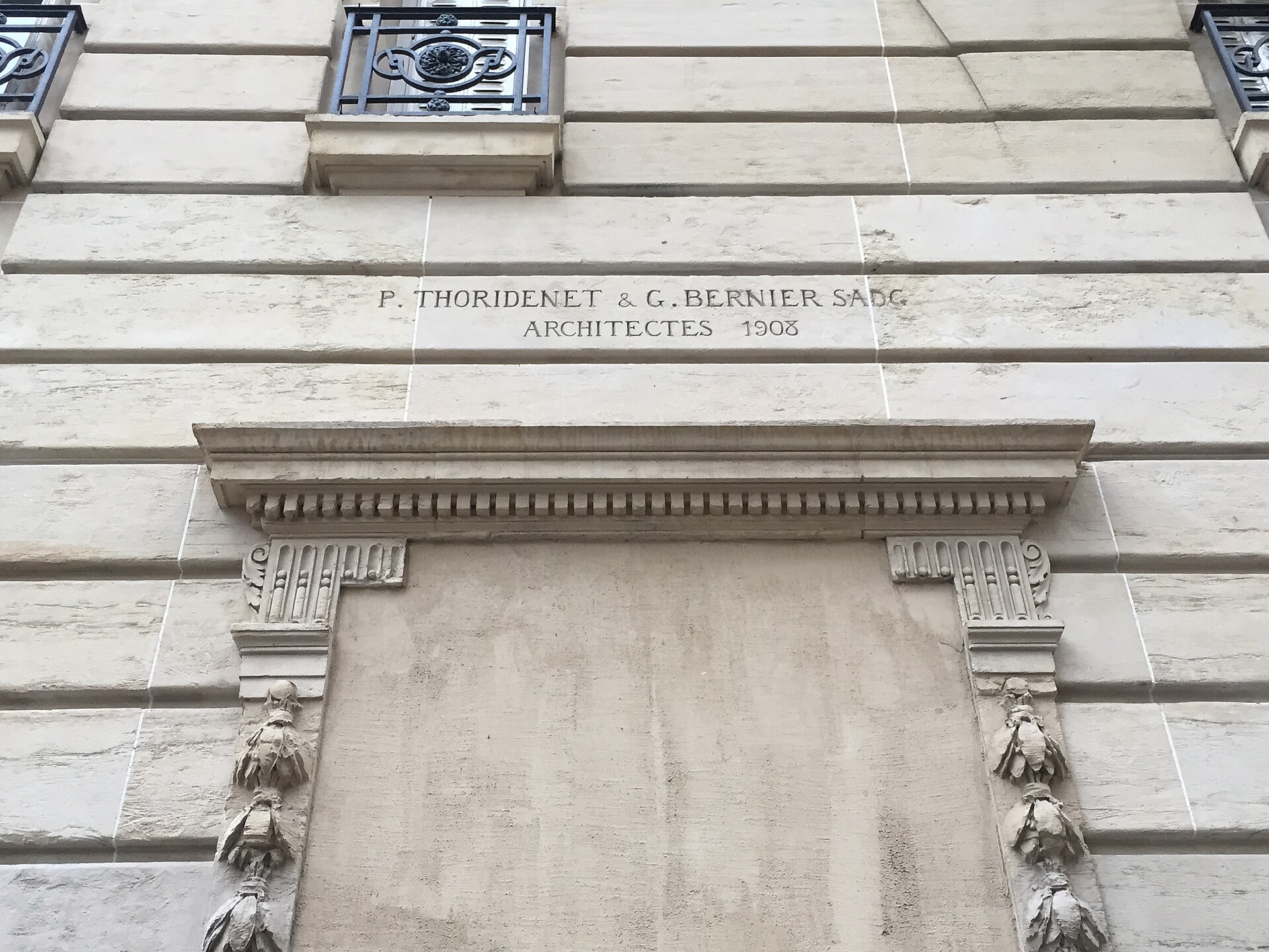 Pale stone building facade with carved architectural ornament and an inscription dated 1908
